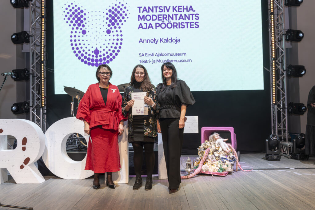 Pildil seisavad Sirje Helme, Annely Kaldoja ja Heidy Purga kõrvuti laval, Kaldoja hoiab käes heledat diplomit. Nende selja taga ekraanil on kirjas "TANTSIV KEHA. MODERNTANTS AJA PÖÖRISTES" / Annely Kaldoja / SA Eesti Ajaloomuuseum / Teatri- ja Muusikamuuseum".