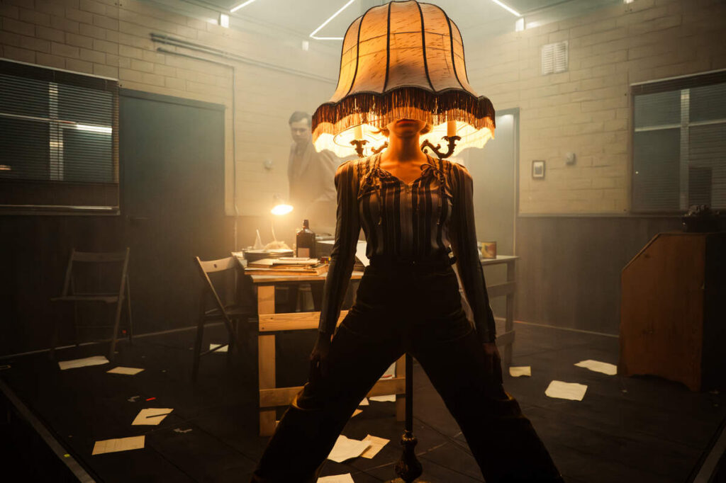 Pildi keskel seisab harkis jalgadega naine, kellel on peas suur antiikne lambikuppel. Taustal on näha lauda, toole ja põrandale kukkunud pabereid.