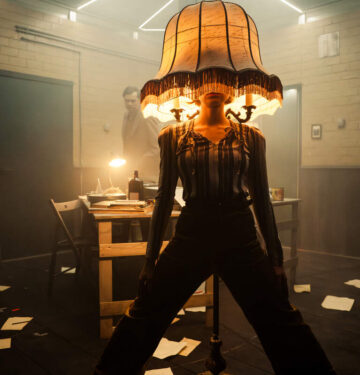 Pildi keskel seisab harkis jalgadega naine, kellel on peas suur antiikne lambikuppel. Taustal on näha lauda, toole ja põrandale kukkunud pabereid.