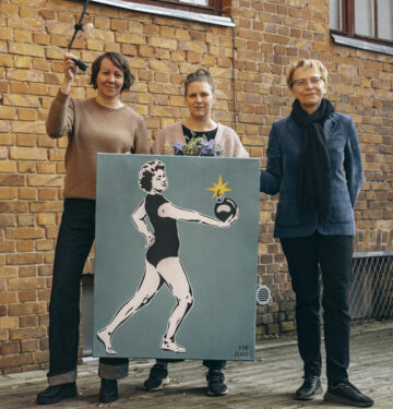 Pildil seisavad Hedi-Liis Toome, Maarja Mänd ja Anneli Saro Tartu Uue Teatri hoovis punase tellisseina taustal. Mänd hoiab käes kunstiteost, mis kujutab naist, kes hoiab käes lõhkevat pommi.