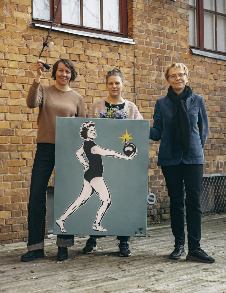 Pildil seisavad Hedi-Liis Toome, Maarja Mänd ja Anneli Saro Tartu Uue Teatri hoovis punase tellisseina taustal. Mänd hoiab käes kunstiteost, mis kujutab naist, kes hoiab käes lõhkevat pommi.