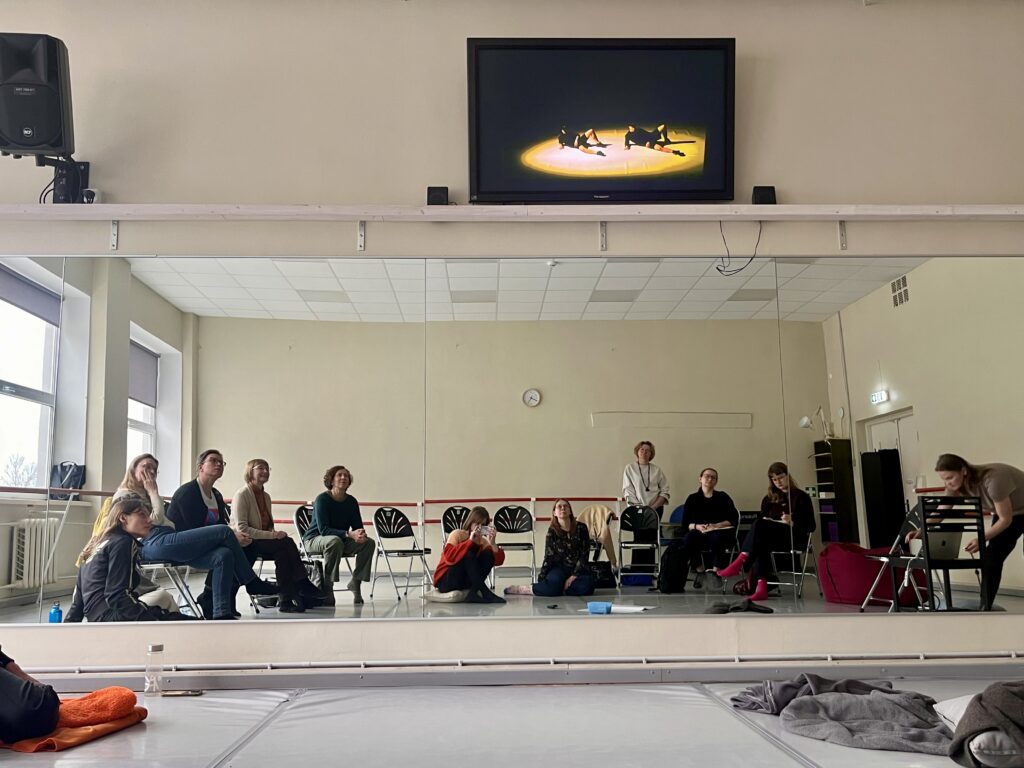 Pildil on näha heledate seinte ja põrandaga tantsusaali, mille seinal oleva peegli peegeldusest paistab kümmekond toolidel ja põrandal istuvat inimest.