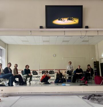 Pildil on näha heledate seinte ja põrandaga tantsusaali, mille seinal oleva peegli peegeldusest paistab kümmekond toolidel ja põrandal istuvat inimest.