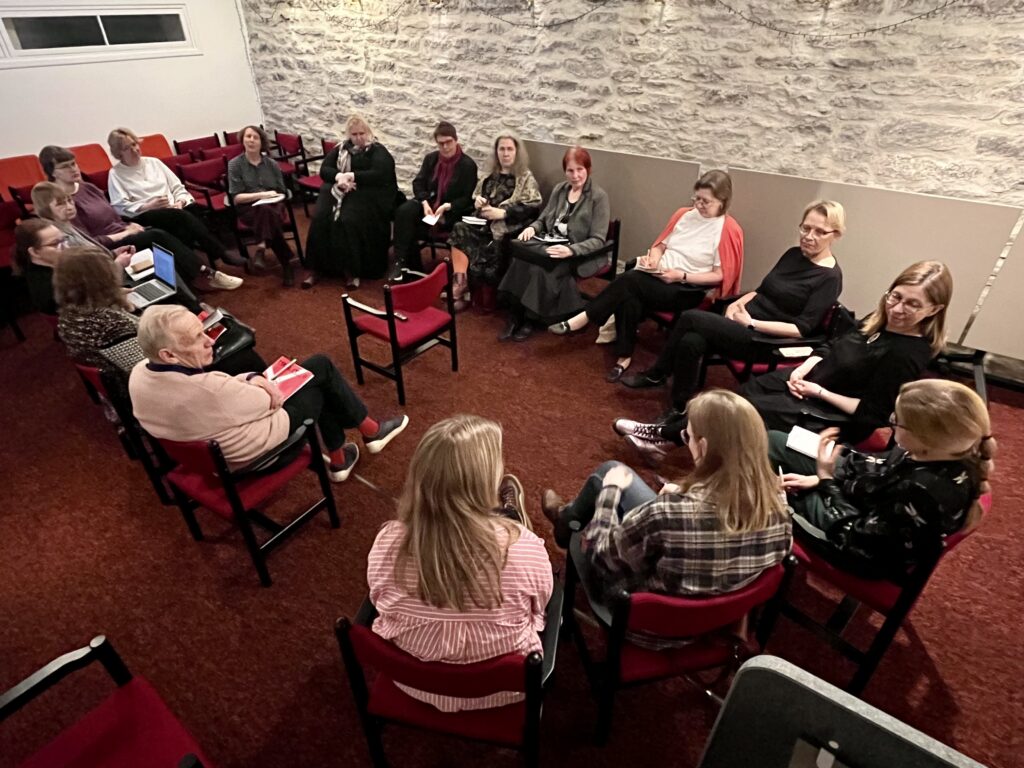 Pildil on punase vaiba ja heleda kiviseinaga saal, kus istuvad ringis toolidel 17 inimest.