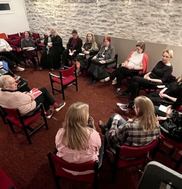 Pildil on punase vaiba ja heleda kiviseinaga saal, kus istuvad ringis toolidel 17 inimest.